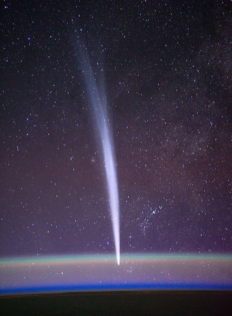 the white streak of a comet stretches downward as it dives below the curved layer of Earth's atmosphere, bulging out with wind as the starry sky of space shines above.