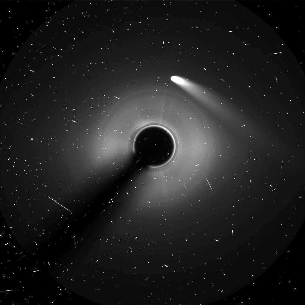 the white streak of a comet extending downward straight from the center, near a black circle surrounded in hazy white, against the starry black of space.