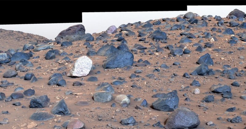 A rocky Martian landscape with various sizes and shapes of blue-toned boulders scattered across a reddish-brown sandy terrain. The earth rises slightly in the background under a pale sky.